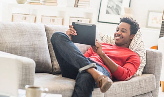 A smiling young man reclines on a sofa and reviews information on a tablet.